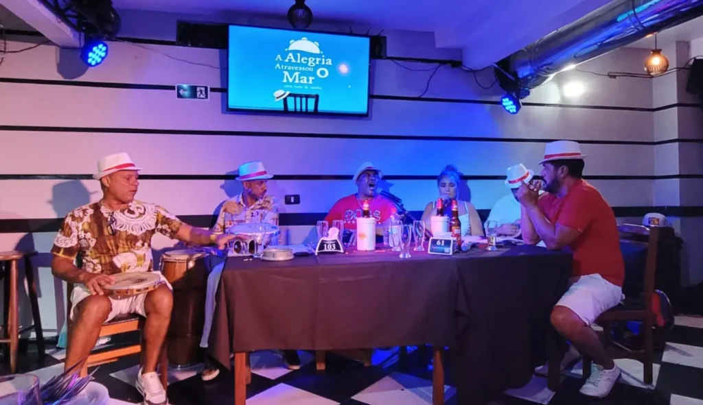 Agenda cultural Santos: cinema, teatro e exposições: Espetáculo musical A Alegria Atravessou o Mar – Uma roda de samba com músicos cantando e tocando instrumentos em um ambiente de evento, com telão ao fundo.