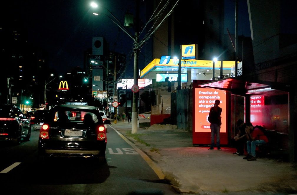 Noite em Campinas, na rua com semáforos e ônibus/parada iluminada. Em destaque, um abrigo virtual “Amigo” em ponto de ônibus com mensagem de apoio e acionamento de serviços policiais para mulheres no Dia das mulheres Campinas.