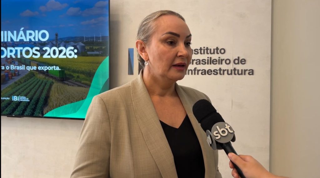 Deputada Daniela Reinehr em entrevista, em evento sobre investimento em infraestrutura, com microfone da SBT e painel institucional ao fundo.