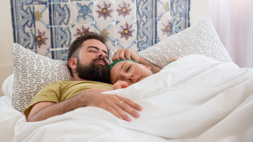 Pessoa demonstrando dificuldade para dormir devido ao ronco ou movimentação do parceiro ao lado, evidenciando os desafios de dividir a mesma cama.