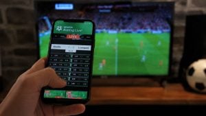 Pessoa utilizando smartphone para realizar apostas esportivas online com elementos visuais da Copa do Mundo 2026 ao fundo.