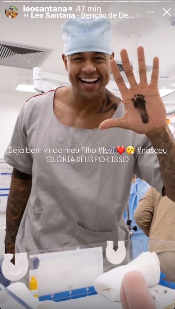 Léo Santana vestindo pijama cirúrgico e exibindo o carimbo do pé do recém-nascido Levi em sua mão, logo após o parto em Salvador.