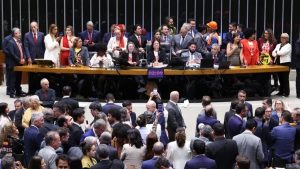 Reunião em plenário com diversas pessoas em volta de uma mesa longa, políticos e autoridades sentados e em pé durante sessão, em ambiente interno