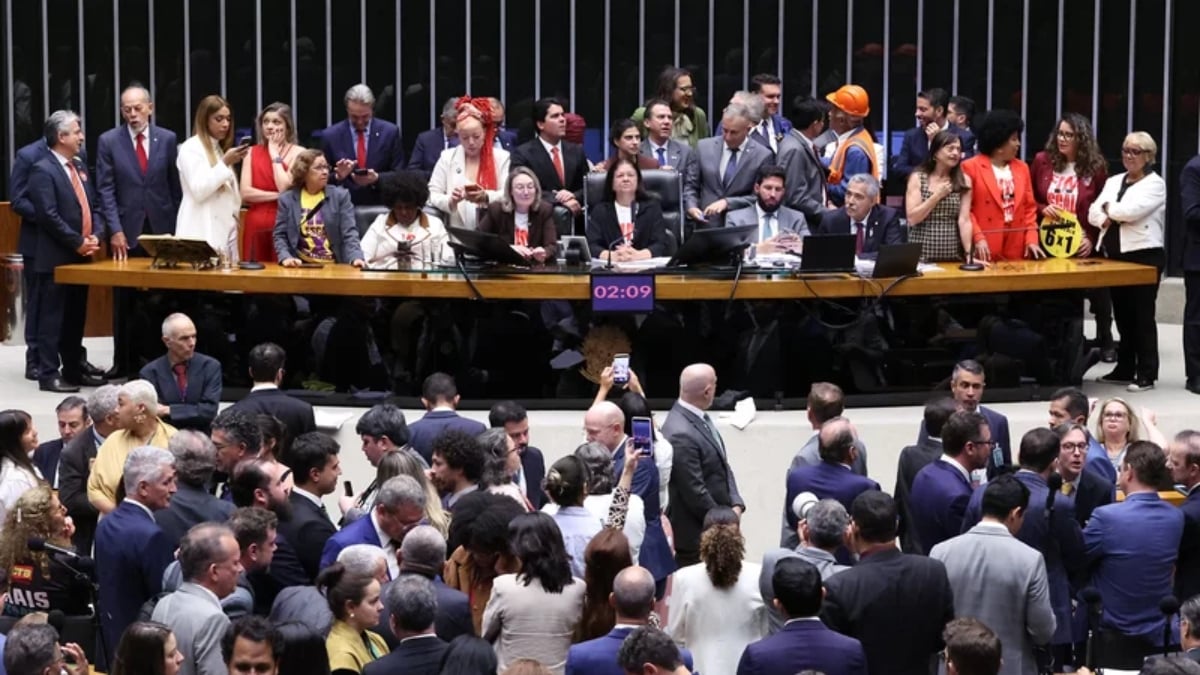Reunião em plenário com diversas pessoas em volta de uma mesa longa, políticos e autoridades sentados e em pé durante sessão, em ambiente interno