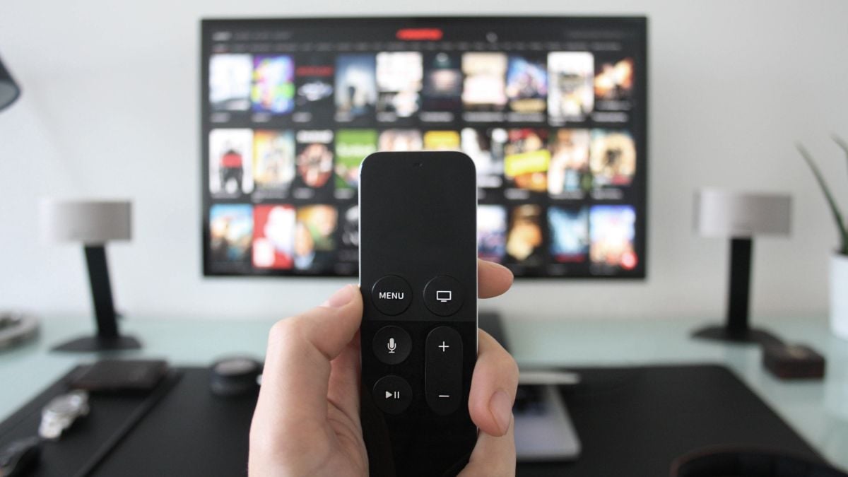 Pessoa segurando um controle remoto em frente a uma TV com várias miniaturas de filmes e séries na tela, destacando recomendações no streaming para produções LGBTQIA+
