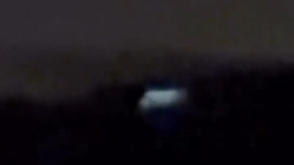 Objeto voador não identificado emitindo luz intensa sobre a linha das árvores em área de mata fechada durante a noite.