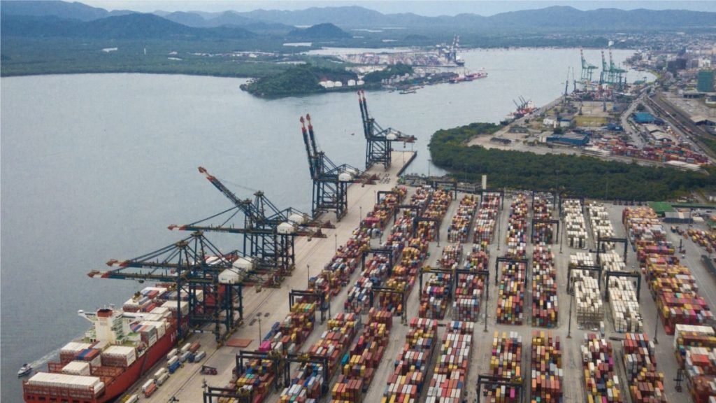 Porto de Santos com vista aérea do cais e filas de contêineres no terminal, navios e atividades portuárias ao lado do canal em cenário urbano costeiro.