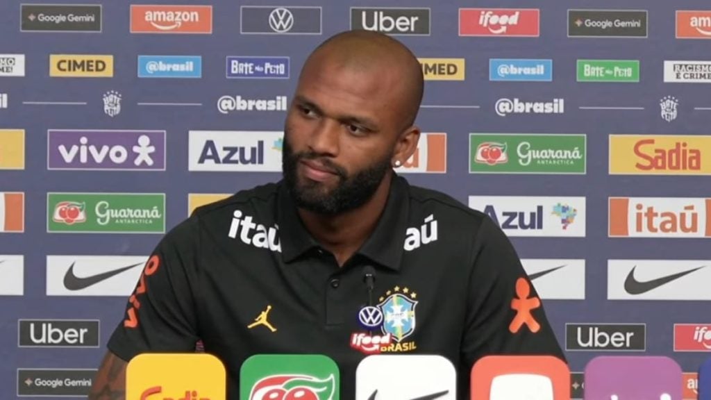 Igor Thiago responde seriamente a uma pergunta na sala de imprensa enquanto Endrick e Rayan participam da dinâmica como jornalistas no centro de treinamentos.