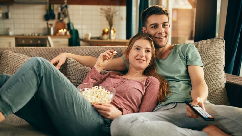 Casal abraçado no sofá assistindo a um filme romântico na televisão, representando uma noite de cinema em casa para o Dia dos Namorados.