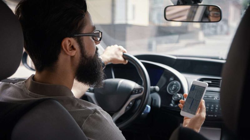 Homem ao volante mexendo no celular representando o número de infrações cometidas pelo uso de celular ao volante.