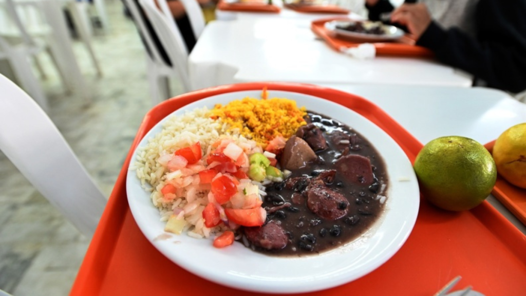 Refeição do programa Bom Prato com cardápio especial inspirado na Copa do Mundo, contendo prato principal balanceado, arroz, feijão e fruta de sobremesa.