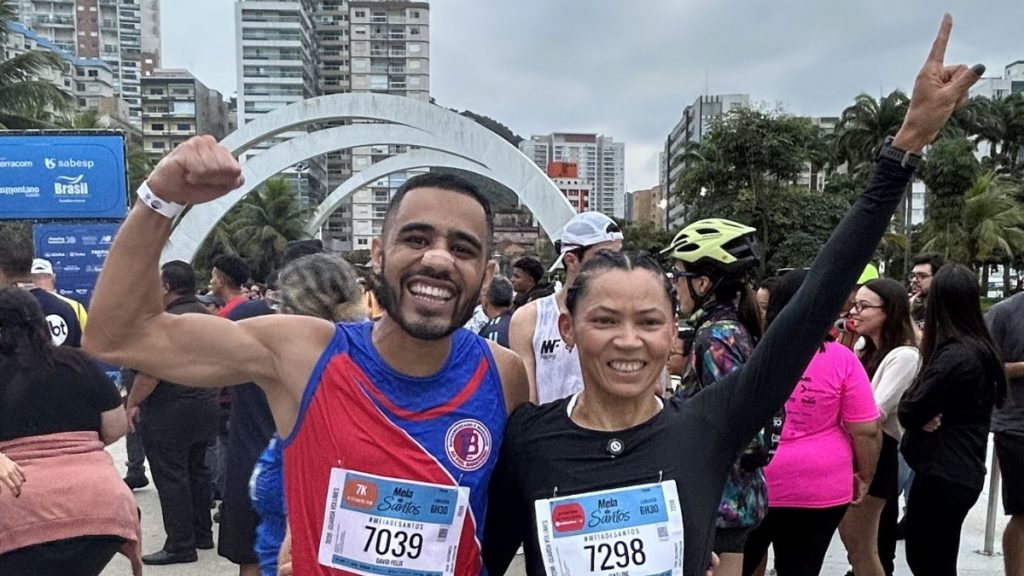 Meia de Santos: Pessoas em corrida urbana comemoram a chegada ao erguer as mãos com sorriso, usando camisetas e luvas de corrida e segurando placas de identificação com números, em meio a prédios e público ao fundo.