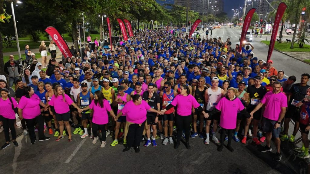 Meia de Santos em destaque na cena de uma corrida lotada à noite, com centenas de participantes usando camisetas e medalhas, reunidos em pista urbana iluminada