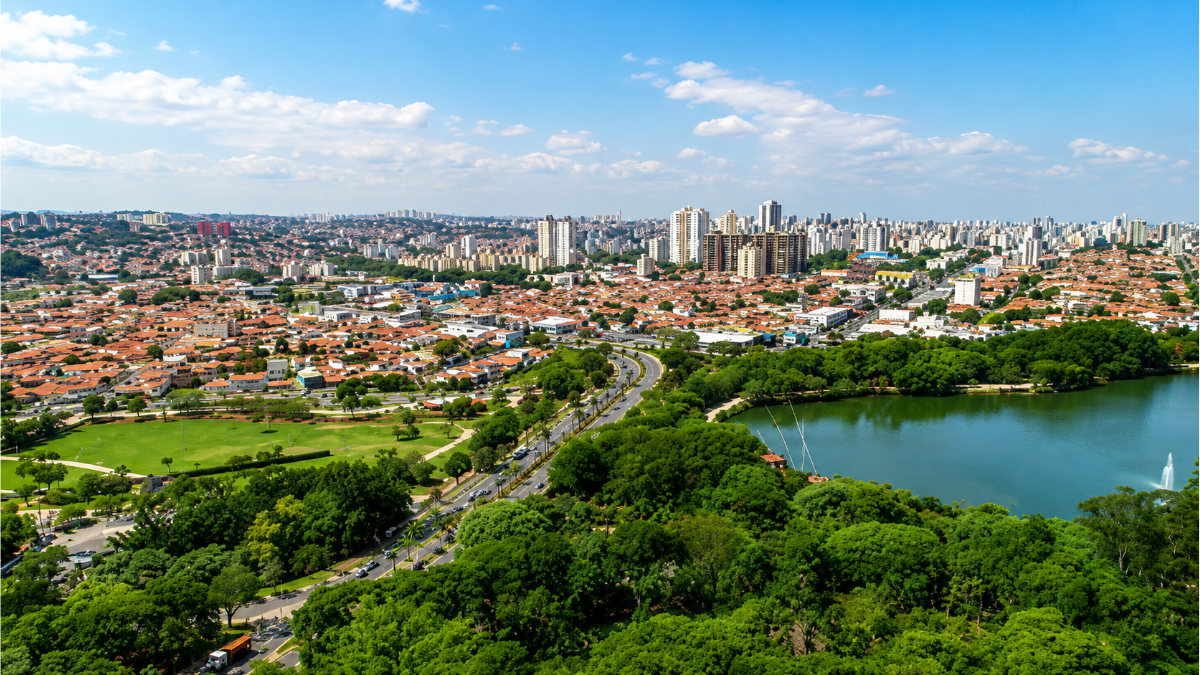 Vista aérea de Campinas, com bairros e um grande parque às margens de um lago, mostrando a cidade ao fundo em dia ensolarado; cenário urbano que ilustra a queda nos roubos na região de Campinas.