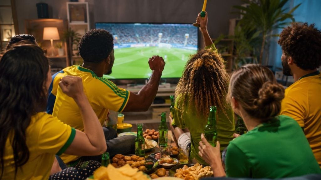 Grupo de pessoas vestidas com amarelo e verde assistindo ao jogo da seleção brasileira na TV durante a Copa, com comidas e bebidas sobre a mesa e clima de torcida em horário comercial.
