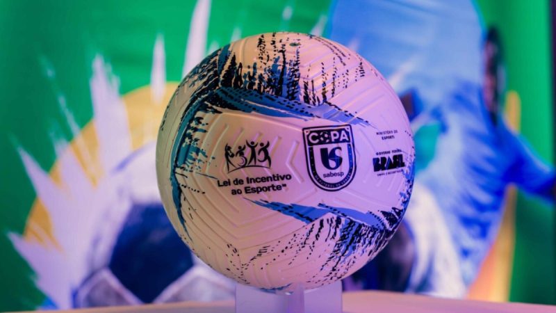 Bola de futebol estampada com selos e logos, em destaque no centro da imagem, em um cenário colorido verde e azul que remete à VTV Copa Sabesp de Futebol de Várzea, perto de evento esportivo