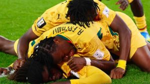 Torcida e jogadores celebram em campo com camisas amarelas da Gana, em um momento de comemoração após uma vitória, com atletas comemorando e abraçando em meio a outro jogador ao chão.