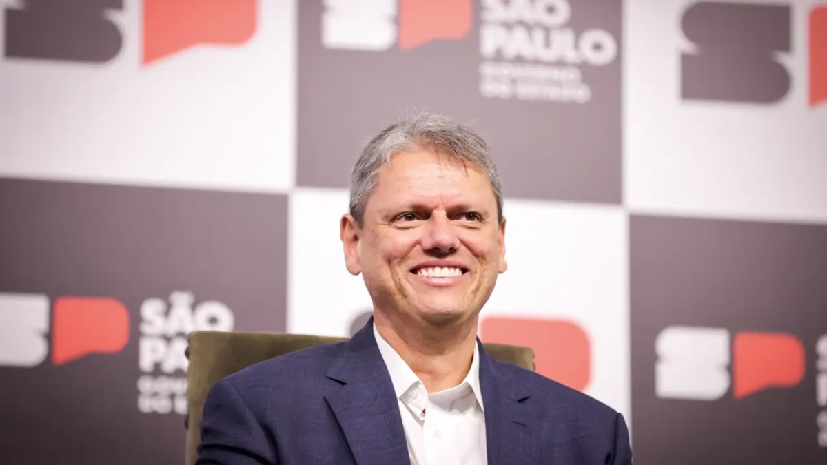 Tarcísio de Freitas sorrindo em evento, usando terno azul e camisa clara, representando sua visita a sede da VTV e participação no VTV da Gente