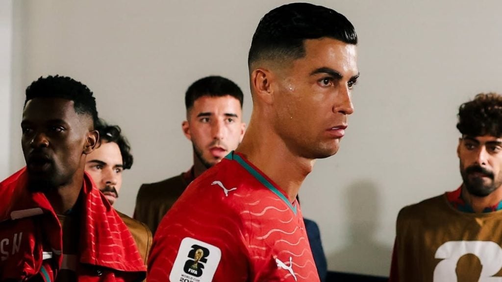 Cristiano Ronaldo em destaque, usando camisa vermelha, enquanto ao fundo aparecem outros jogadores em um ambiente interno, com clima de coletiva ou jogo.