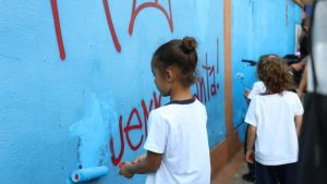 Criança e estudantes observados na escola durante aula, com profissional ao fundo, em cenário de sala com quadro e parede azul, destacando a identificação precoce de estudantes com altas habilidades.