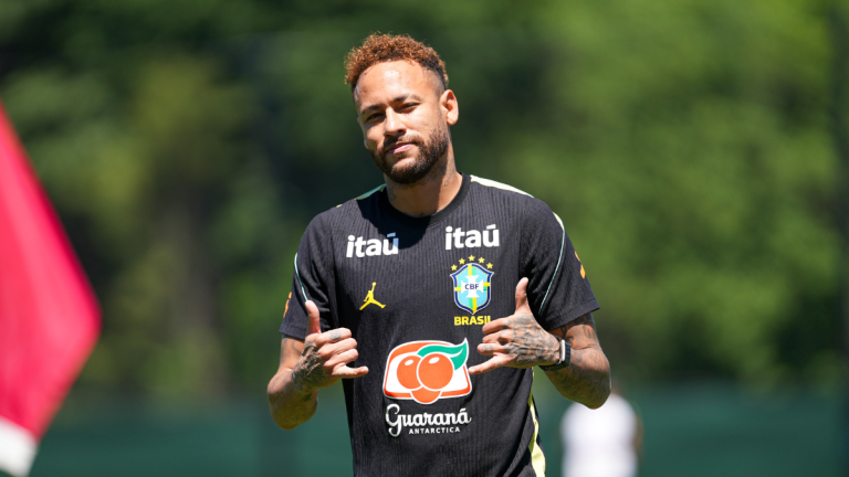 Neymar durante treino da Seleção Brasileira em campo, com a camisa da equipe e patrocinadores visíveis, falando ao lado de bandeira, em preparação para o jogo contra o Haiti.
