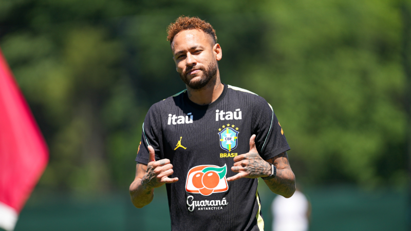 Neymar durante treino da Seleção Brasileira em campo, com a camisa da equipe e patrocinadores visíveis, falando ao lado de bandeira, em preparação para o jogo contra o Haiti.