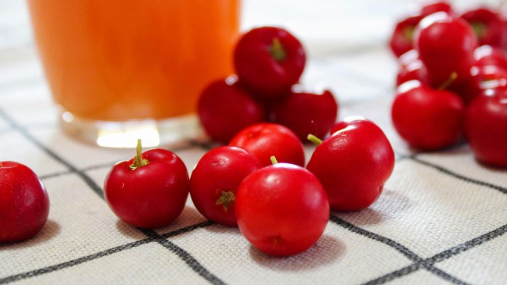 Acerola fresca em destaque, com suco ao fundo em copo, mostrando os frutos vermelhos sobre uma superfície clara, reforçando a alta concentração de vitamina C.