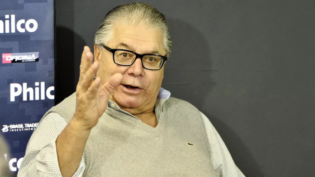 Presidente do Conselho Deliberativo da Ponte Preta SAF, José Armando Abdalla Junior, em entrevista, segurando a mão no rosto durante evento com fundo de mídia ao lado de logotipo.