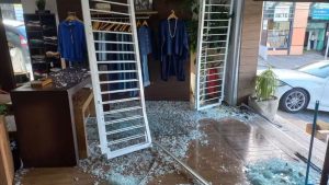 Loja de roupas após acidente em Vinhedo: carro invadiu a fachada, deixou vidros e objetos no chão e atingiu a entrada com grades e portas danificadas; ninguém ficou ferido e condutora assumiu os prejuízos.