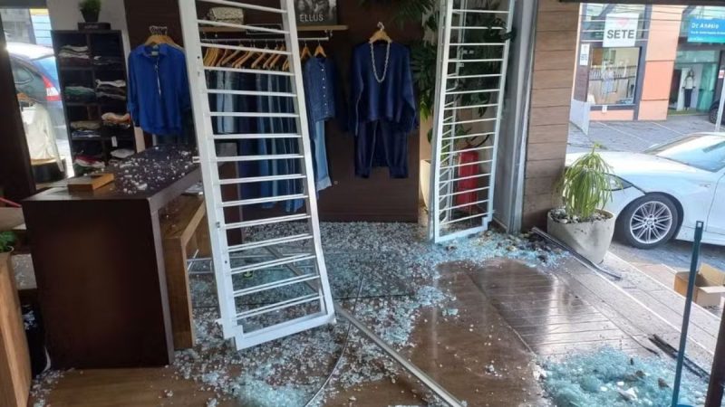 Loja de roupas após acidente em Vinhedo: carro invadiu a fachada, deixou vidros e objetos no chão e atingiu a entrada com grades e portas danificadas; ninguém ficou ferido e condutora assumiu os prejuízos.