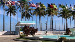 Praia com piscina ao ar livre e bandeiras em frente a um hotel na Baixada Santista, com palmeiras e céu azul, cenário para o jogo do Brasil que abre e fecha na região.