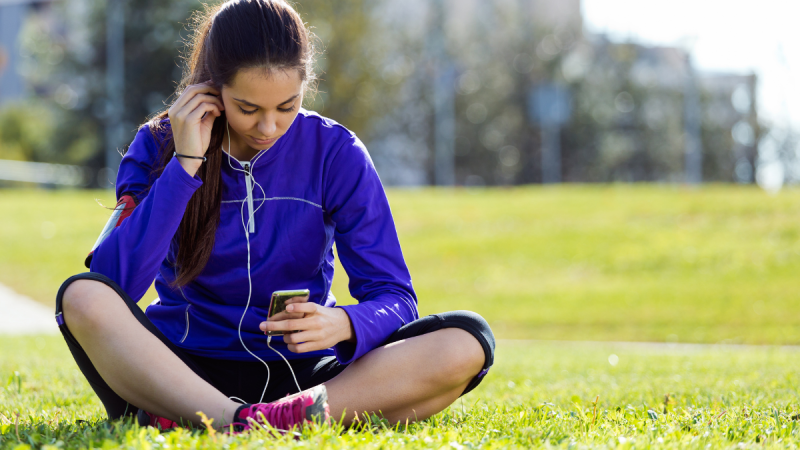 Jovem sentada na grama em um parque ouvindo música pelo celular com fones de ouvido. Imagem que remete às Músicas das edições da Copa, trilhas marcantes do futebol de 1970 a sucessos atuais.