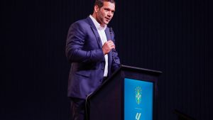 Samir Xaud em treino da Seleção, aparece falando ao microfone em um púlpito durante evento com fundo escuro e emblema na tela ao lado, expressão séria e postura de apresentação