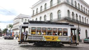 Bonde da Copa em passeio temático na cidade, com passageiros dentro do veículo decorado na rua de paralelepípedos diante de prédio histórico ao fundo.