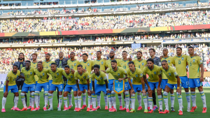 Seleção brasileira em campo durante partida de futebol, com jogadores usando o uniforme amarelo e azul e bandeira/escudo da Copa do Mundo 2026, em estádio lotado.