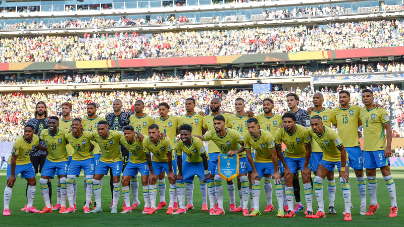 Seleção brasileira em campo durante partida de futebol, com jogadores usando o uniforme amarelo e azul e bandeira/escudo da Copa do Mundo 2026, em estádio lotado.