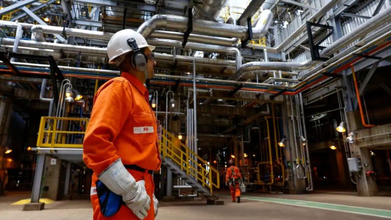 Trabalhador em fábrica usando capacete e uniforme laranja em meio a tubulações industriais, cenário que ilustra a economia brasileira e seus efeitos no emprego e na produção.