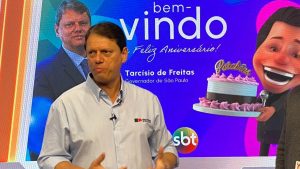Tarcísio de Freitas em entrevista, com o fundo “bem-vindo” e o texto “Tarcísio de Freitas” e “Governador de São Paulo”, enquanto discute a ligação entre Santos e Guarujá prevista até 2030.
