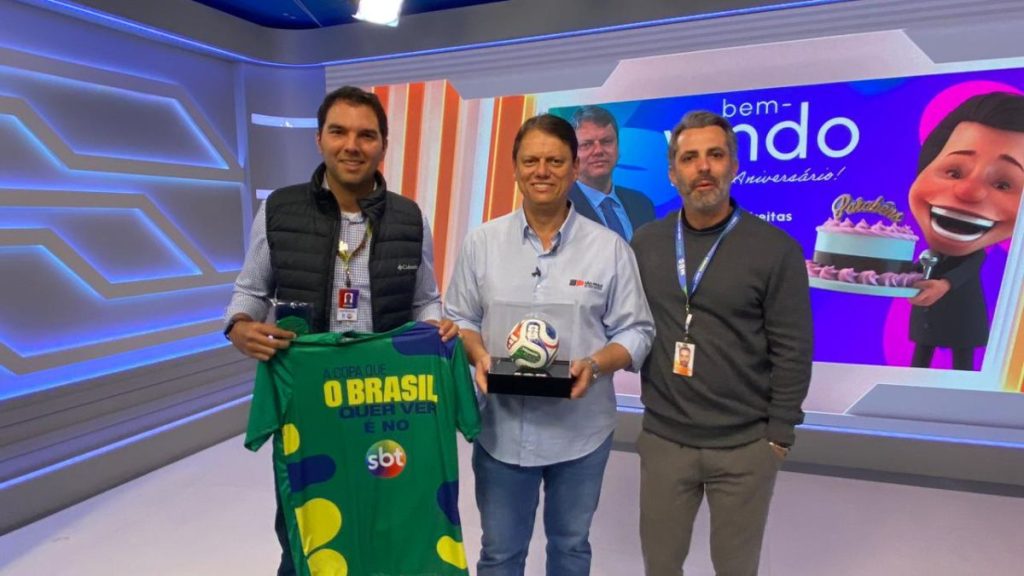 Tarcísio, acompanhado por dois homens em um estúdio, recebe uma bola Trionda oficial da Copa do Mundo de 2026 durante participação no SBT, em evento relacionado ao túnel Santos-Guarujá Tarcísio.