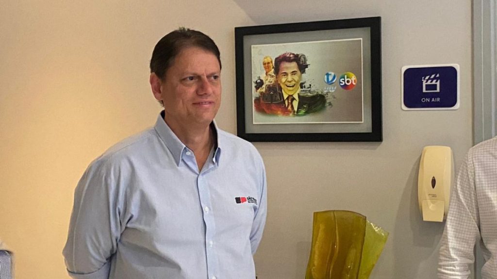 Governador em visita à VTV, em sala interna, falando ao lado de quadro na parede e apontando informações sobre o Túnel Santos-Guarujá Tarcísio e prazos do projeto