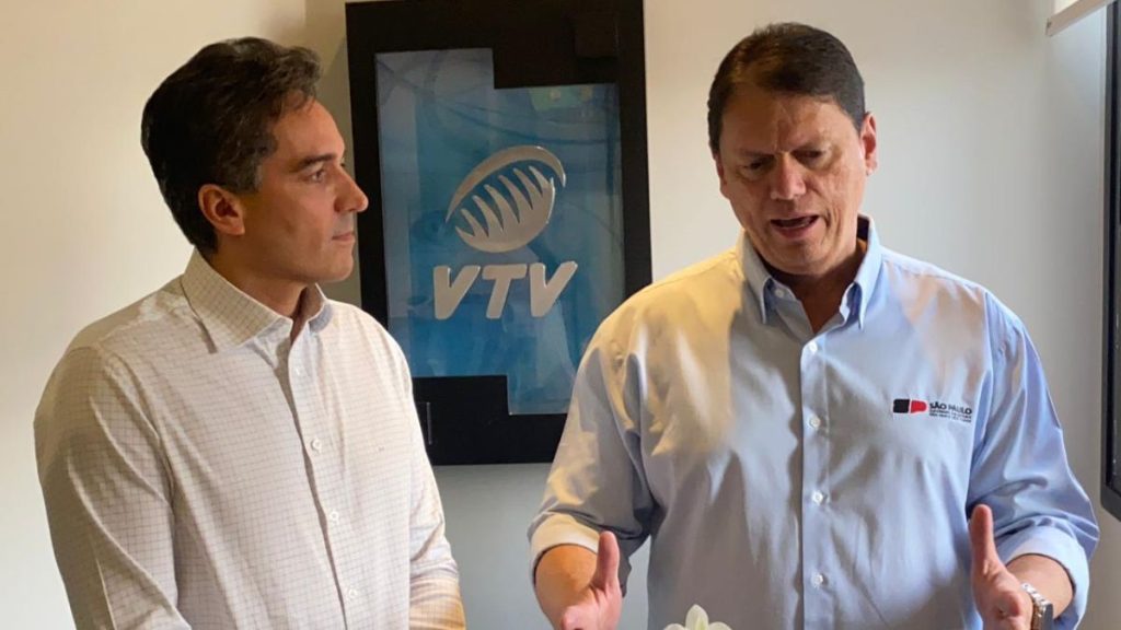 Tarcísio conversa durante entrevista em Santos, ao lado de entrevistador, mencionando investimentos na região. Ao fundo, sinalização da VTV e informações do evento.
