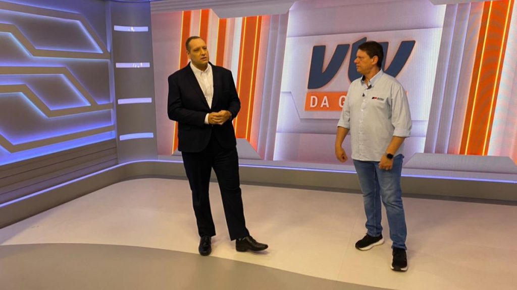 Apresentação em estúdio de TV com Tarcísio ao lado de apresentador, com destaque para o Túnel Santos-Guarujá e projetos de saneamento na Baixada Santista, em cenário com telão ao fundo