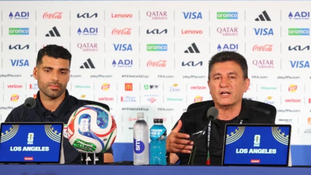 Mehdi Taremi, capitão da seleção do Irã, em coletiva de imprensa após reclamação na FIFA, ao lado do técnico Amir Ghalenoei falando sobre o assunto ao fundo com logotipos e bandeiras de patrocinadores.