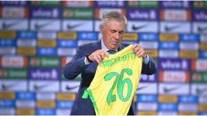 Carlo Ancelotti segura a camisa amarela com o número 26 em um cenário de fundo com bandeiras e patrocinadores, em foto relacionada ao ranking de técnicos mais bem pagos da Copa do Mundo.