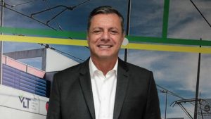 O prefeito de Santos em foto de reportagem, usando terno escuro e camisa branca, em ambiente externo com estrutura e bandeiras ao fundo, enquanto informa afastamento para tratar câncer.
