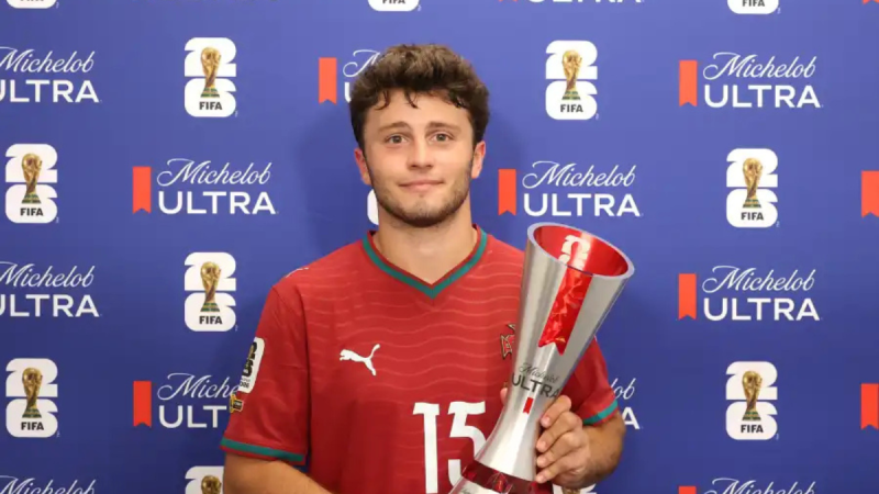João Neves aparece em foto de premiação em um evento de futebol da FIFA, vestindo camisa vermelha e segurando uma grande taça, em frente a um painel azul com o logotipo FIFA e Michelo ULTRA.