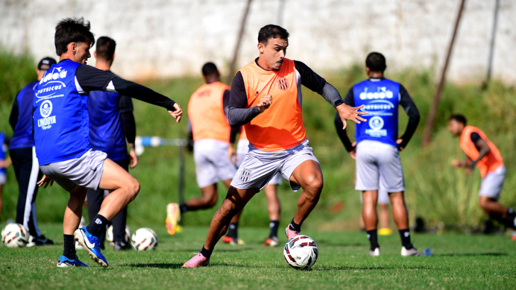 Willian Pottker jogando futebol durante treino da Ponte Preta em campo, com bola entre os jogadores e uniforme azul e laranja na atividade esportiva.