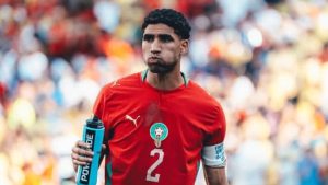 Hakimi, astro do Marrocos, em campo durante partida, usando camisa vermelha da seleção e segurando uma garrafa azul de água. Ele aparece em destaque com o número 2 na camisa.