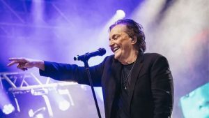 Show Fabio Junior Campinas: cantor Fábio Júnior em palco, sorrindo e apontando para o público, com microfone e iluminação roxa durante apresentação ao vivo.