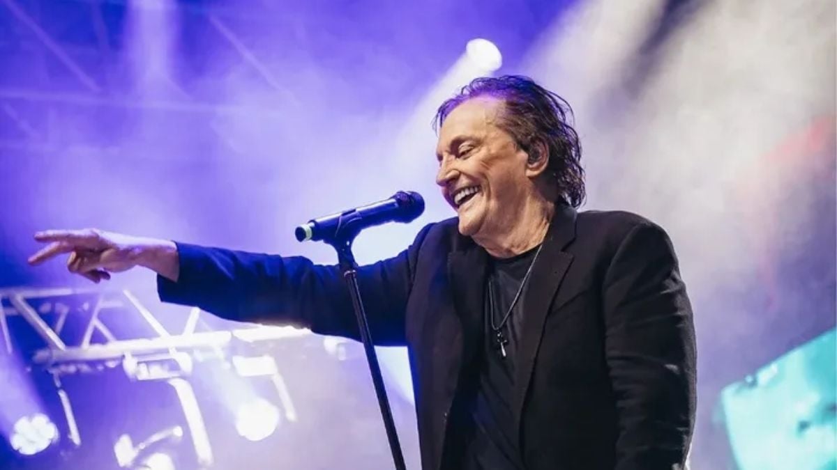 Show Fabio Junior Campinas: cantor Fábio Júnior em palco, sorrindo e apontando para o público, com microfone e iluminação roxa durante apresentação ao vivo.
